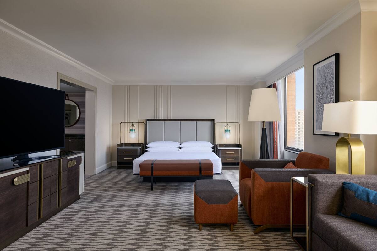 Quarto de hotel com uma cama grande, televisão, duas mesas de cabeceira com luminárias, sofá, banquinho e vista para a cidade pela janela.