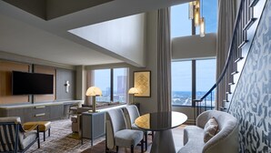 Quarto de hotel moderno com televisão grande, assentos confortáveis, área de refeições e vista para a paisagem urbana.