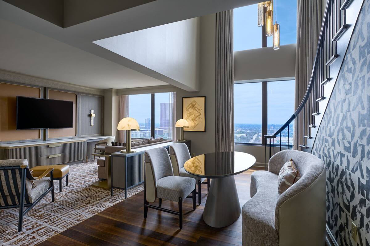 Quarto de hotel moderno com televisão grande, assentos confortáveis, área de refeições e vista para a paisagem urbana.