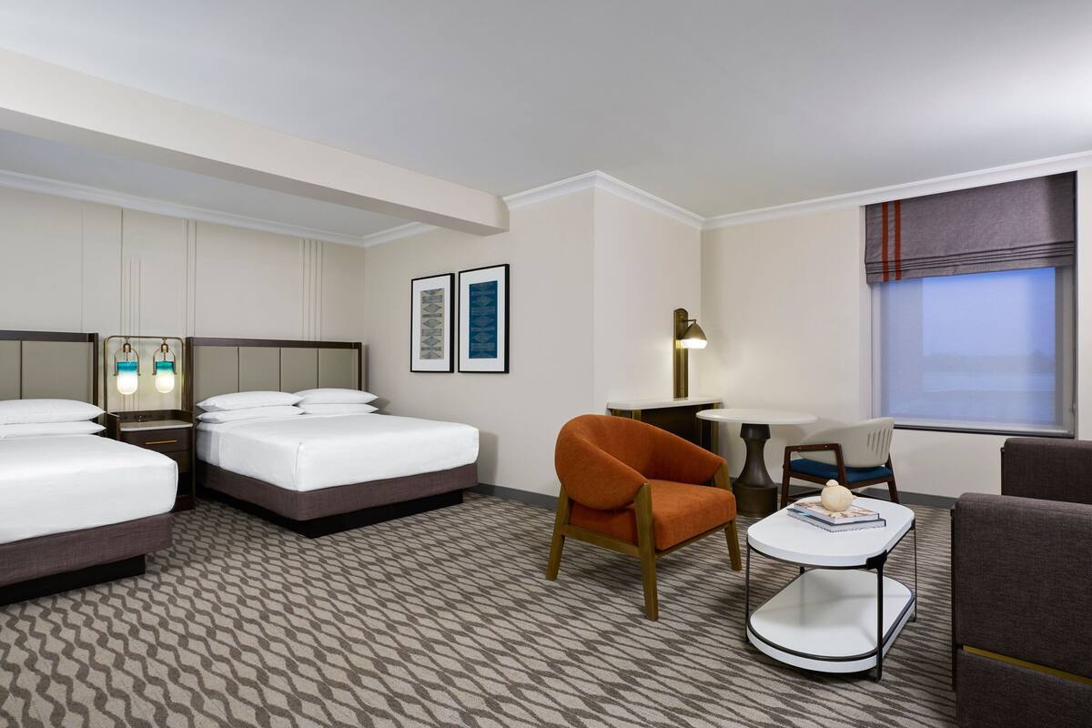 Quarto de hotel com duas camas, uma mesinha, uma cadeira e uma luminária.