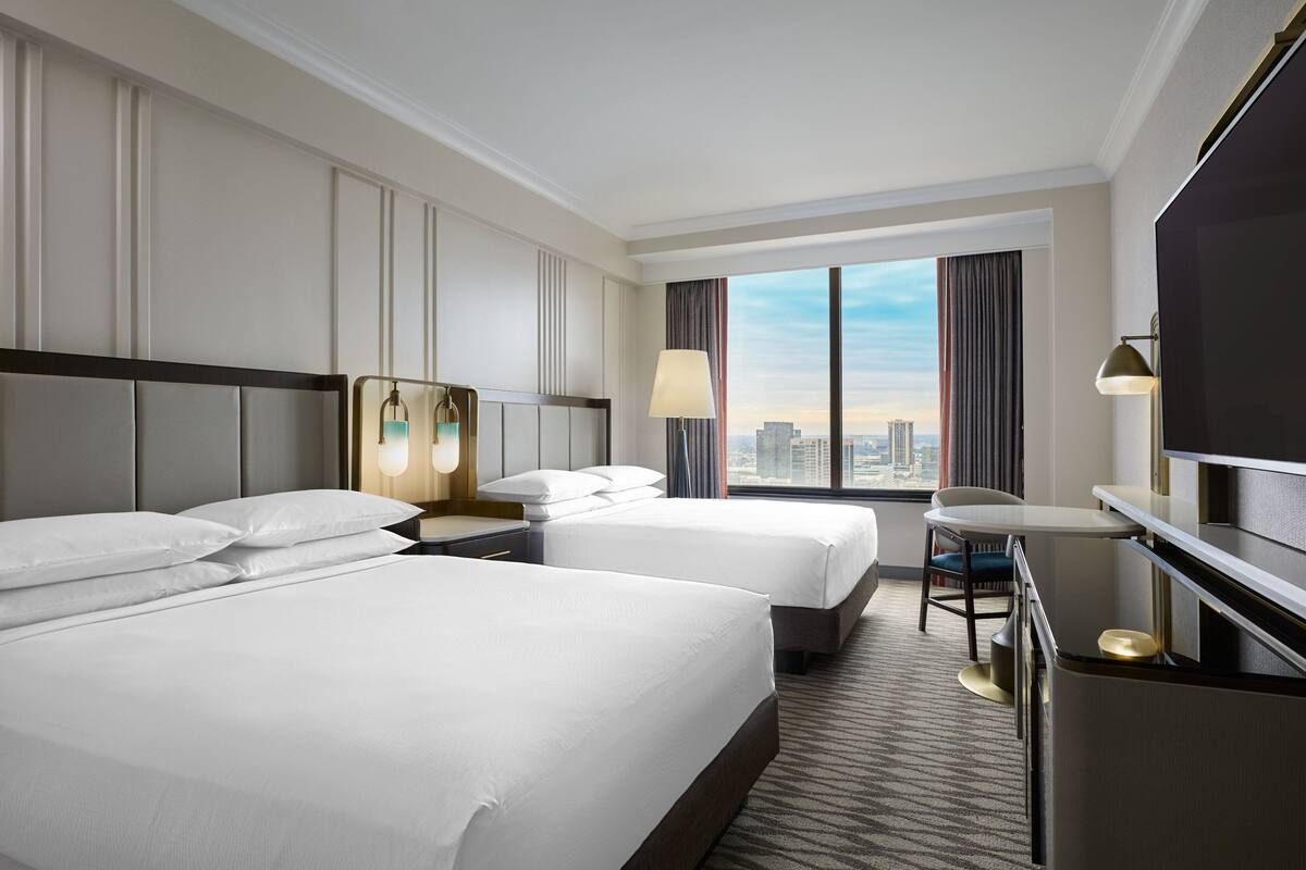 Quarto de hotel com duas camas, uma escrivaninha, uma cadeira, uma televisão e uma janela grande com vista para a cidade.