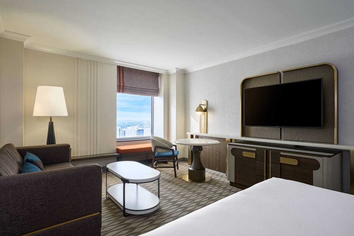 Quarto de hotel com cama, sofá, mesa pequena, cadeira, escrivaninha, televisão e janela com vista para a cidade.