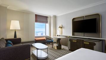A hotel room with a bed, a sofa, a small table, a chair, a desk, a television, and a window with a view of the city.