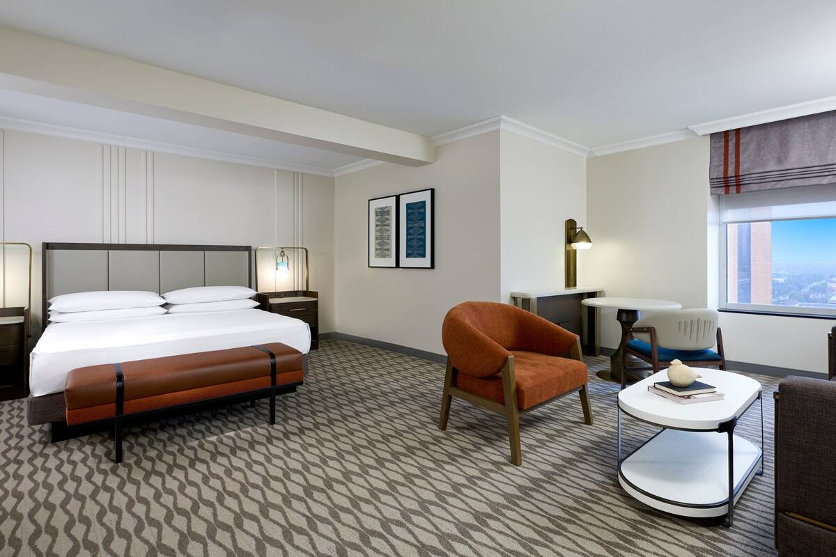 Quarto de hotel com cama, um banco, uma cadeira, uma mesinha e um abajur.