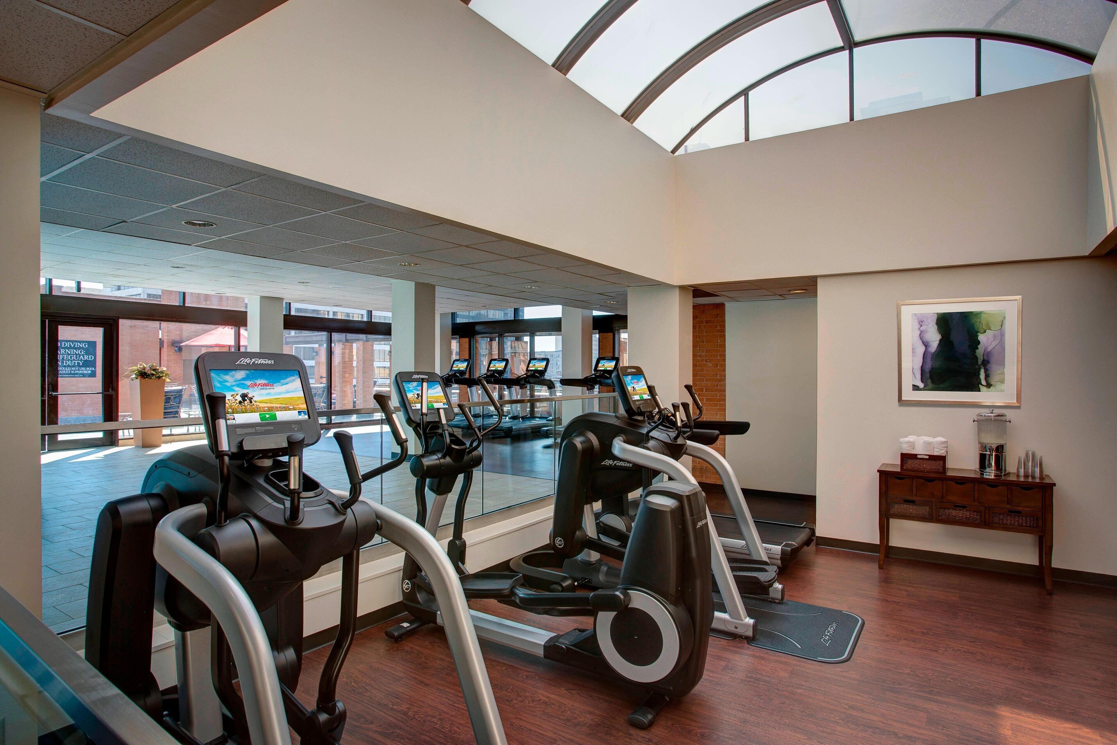 Ein Fitnessraum mit Laufbändern und Crosstrainern, Holzboden, einem großen Fenster und einem Wandbild.