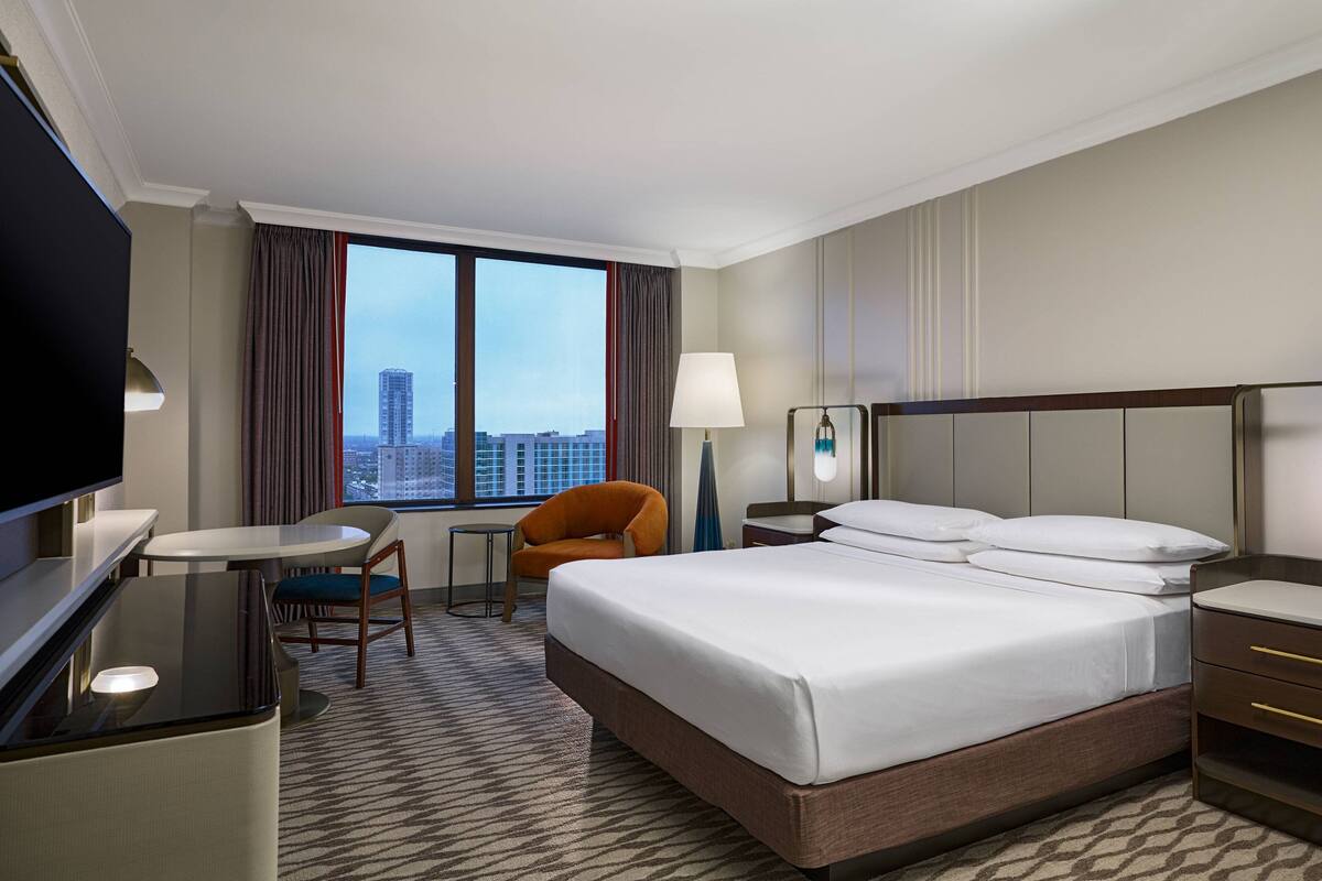 Quarto de hotel com uma cama grande, uma escrivaninha, uma cadeira, televisão e vista para a cidade.