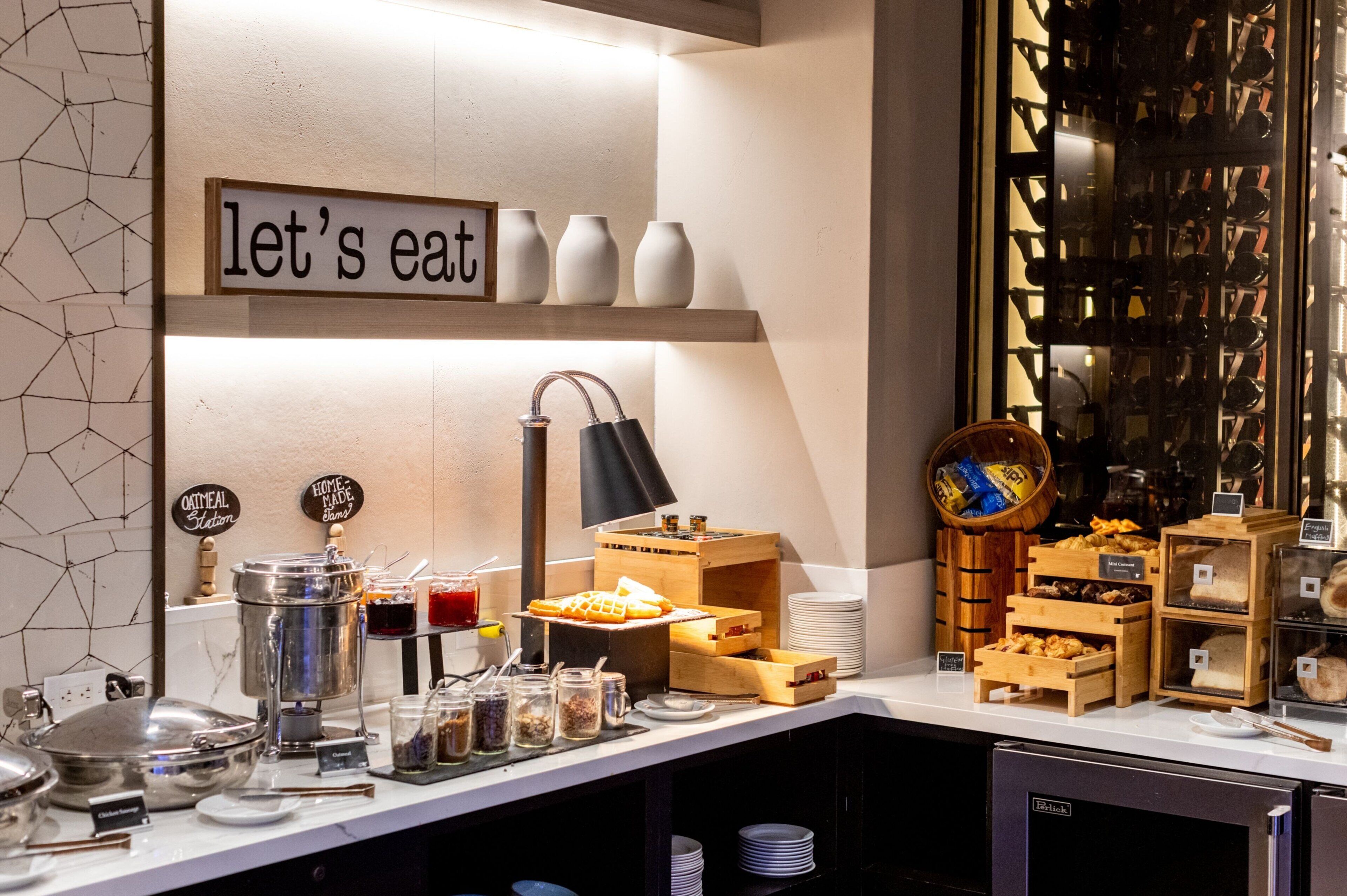 A breakfast buffet with various food items, a sign that says "let's eat", and a coffee machine.