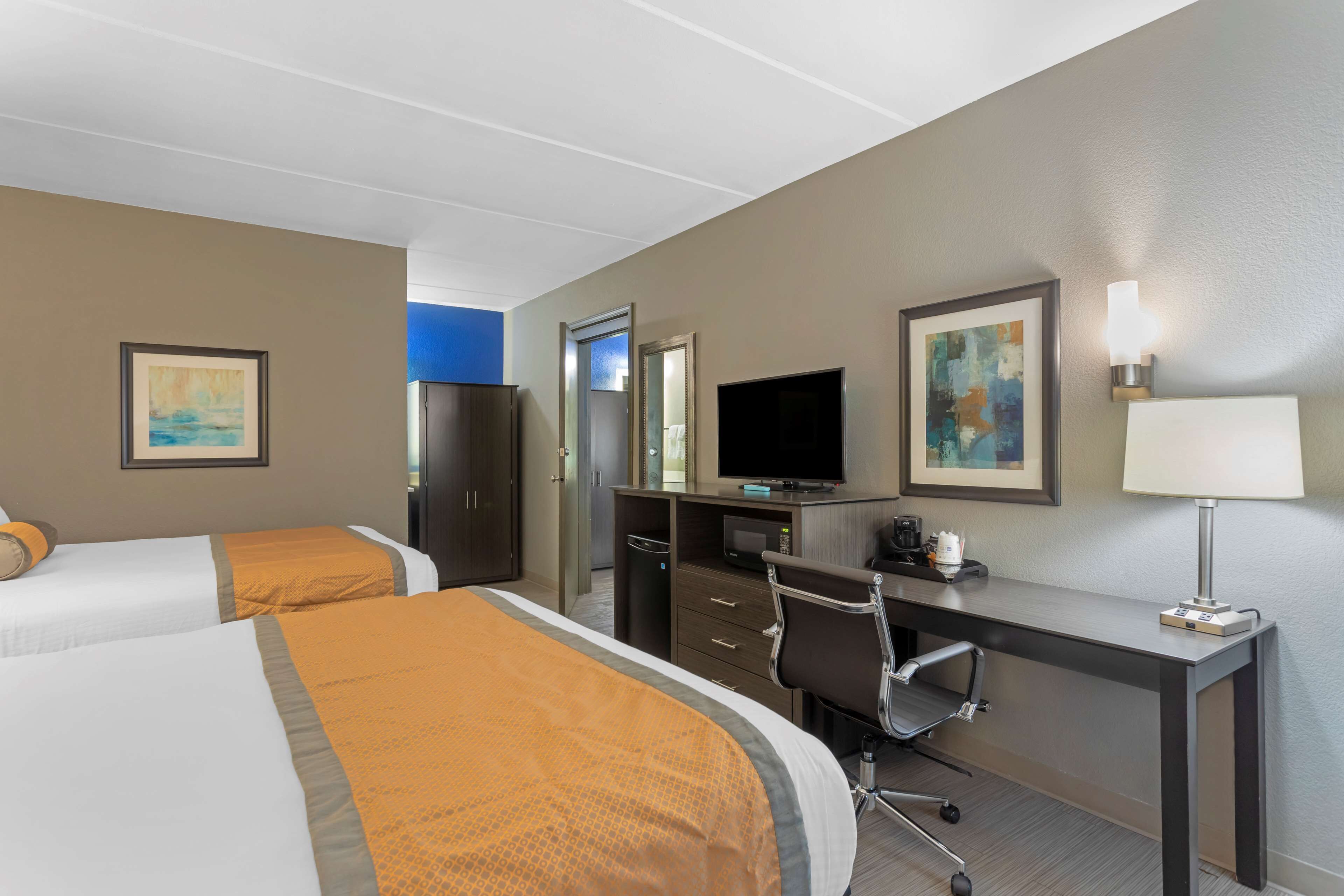 A hotel room with two beds, a desk, a chair, a television, and artwork on the walls.