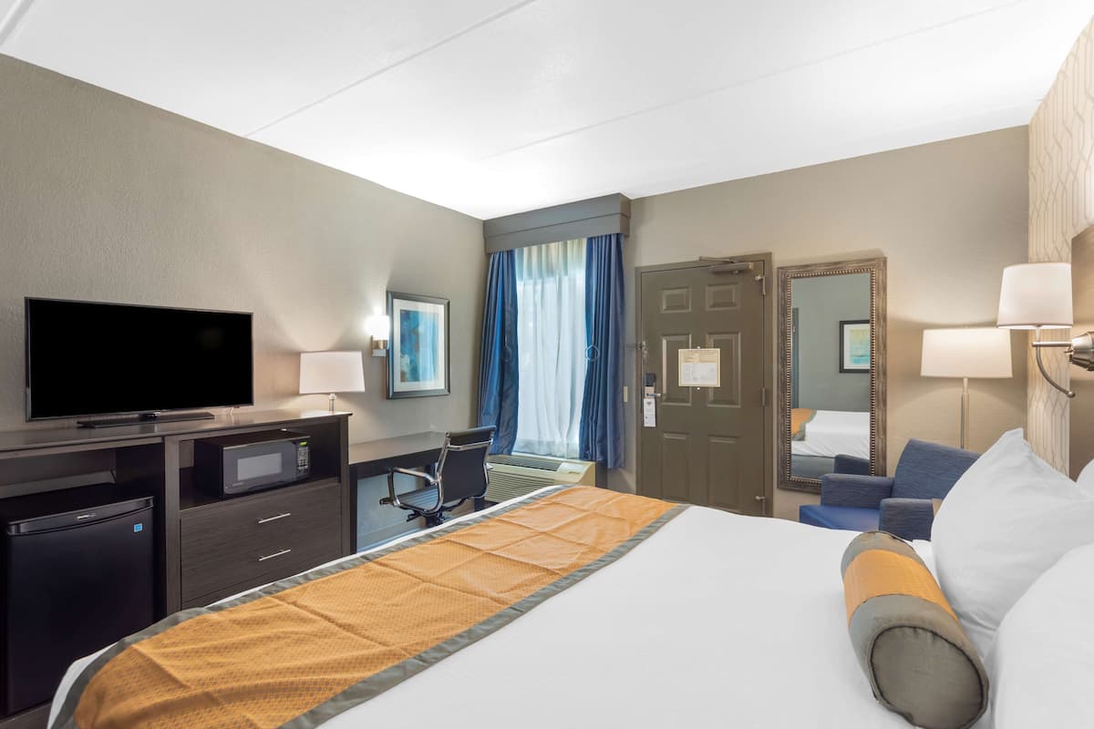A hotel room with a bed, a desk with a flat-screen TV, a microwave, a mini-fridge, a chair, a desk lamp, and a large mirror.