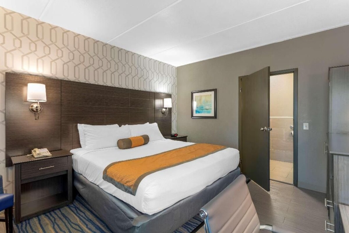 Una habitación de hotel con una cama grande, mesitas de noche, una lámpara, un cuadro y una puerta que da a un baño.