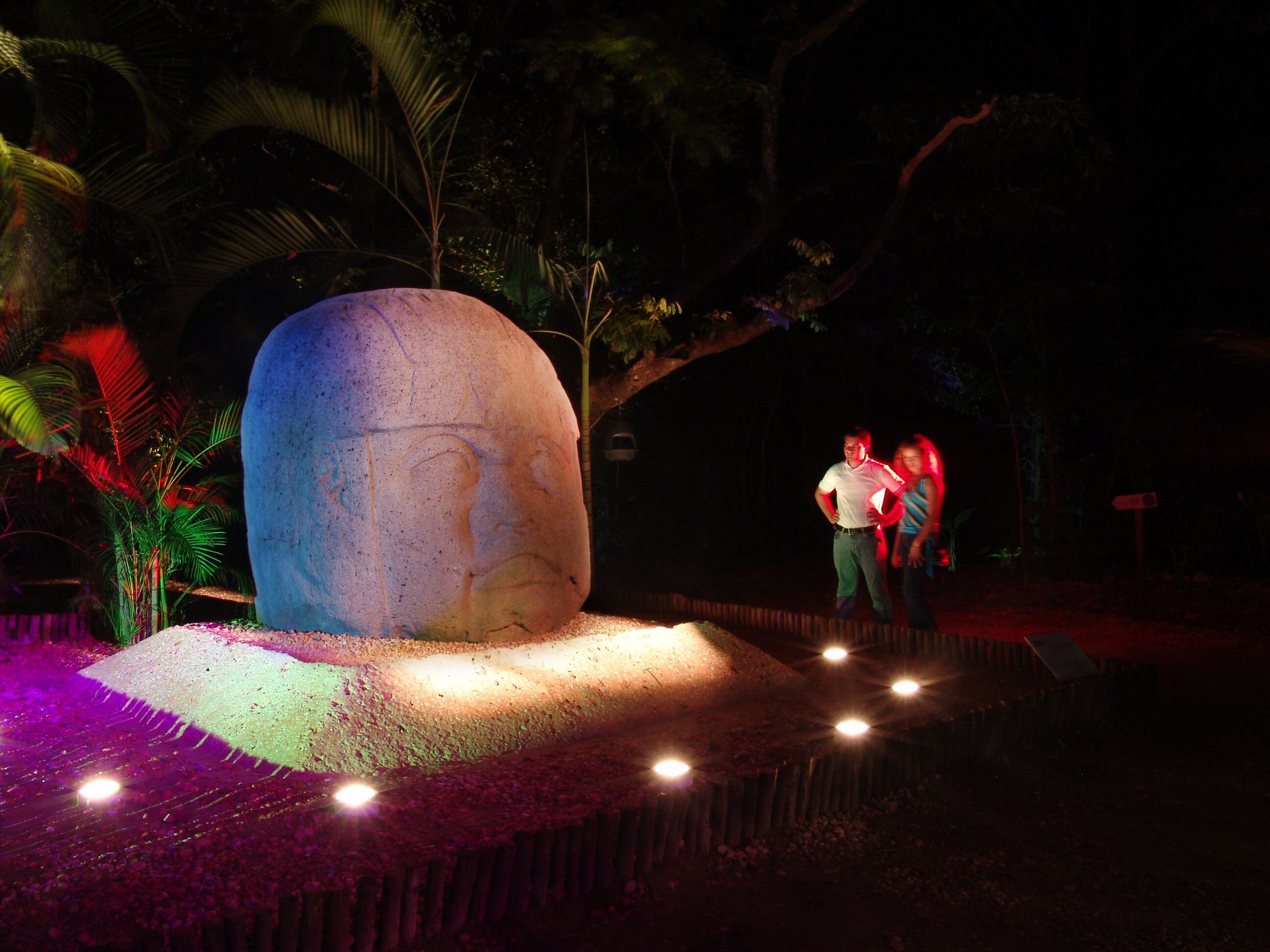 A large stone monument with a carved face, surrounded by palm trees and illuminated by spotlights.