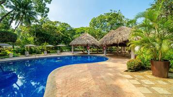 A swimming pool with a tiled deck, surrounded by lush greenery and thatched-roof structures.