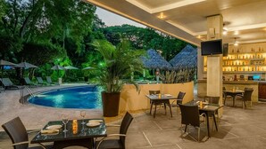 Un espace extérieur avec une piscine, des chaises longues et des parasols, un bar avec des étagères remplies de bouteilles, et un coin repas avec des tables et des chaises.