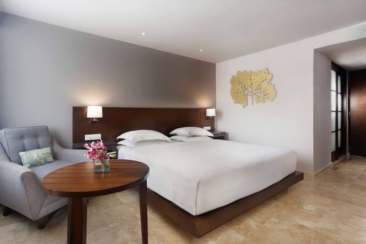 A hotel room with a large bed, a chair, a small table with a vase, and a wall art piece.