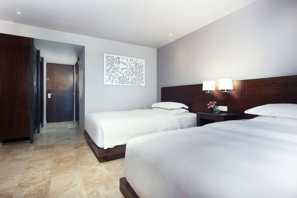 A hotel room with two beds, a wooden headboard, a nightstand with lamps, and a wall art piece.