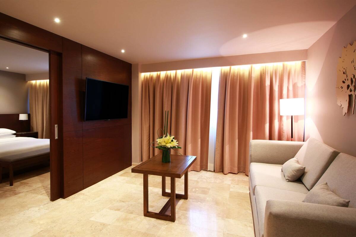 A hotel room with a bed, a TV mounted on the wall, a table with a vase of flowers, a sofa, and curtains.