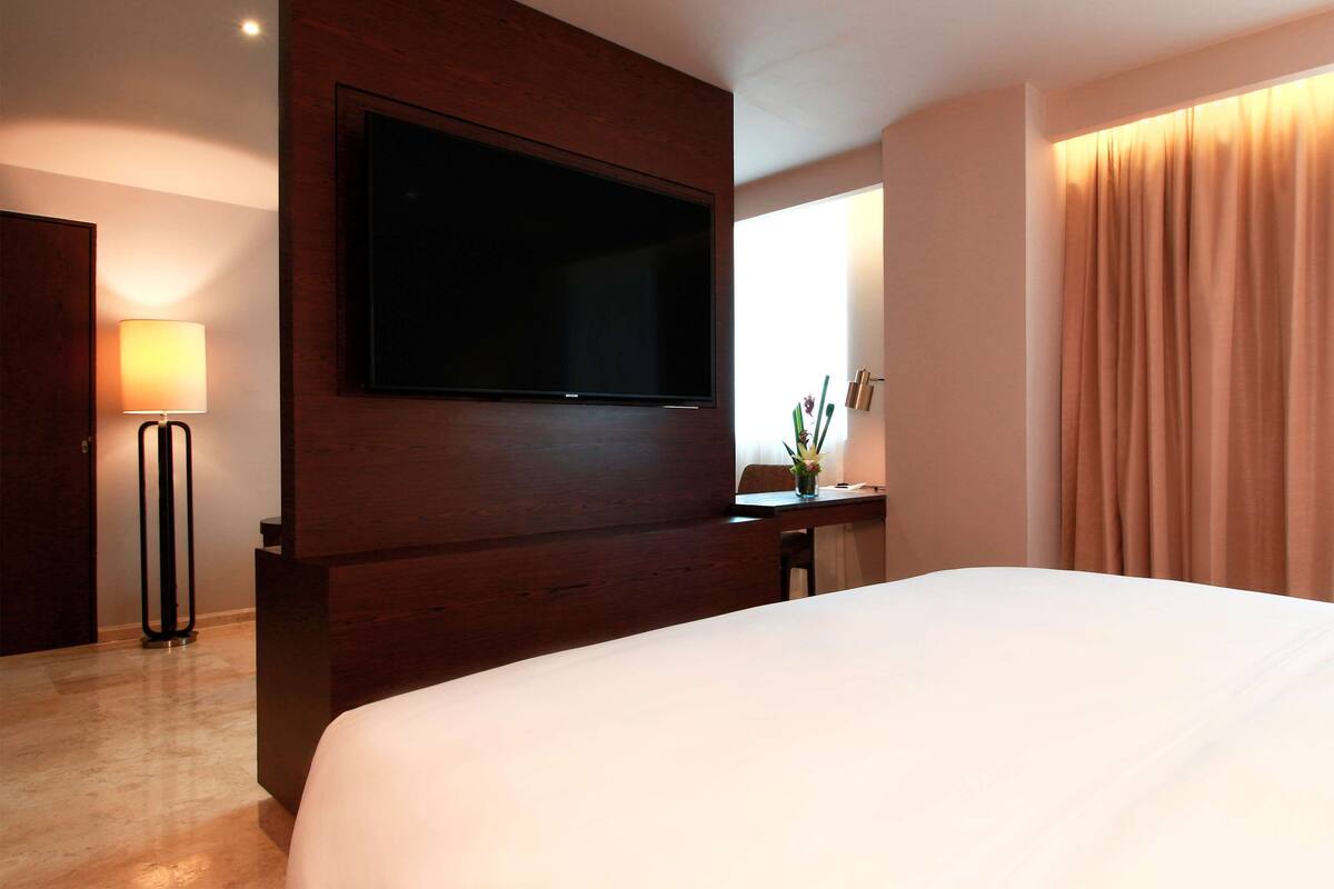 A hotel room with a large bed, a flat-screen TV mounted on the wall, a desk with a lamp, and a window with curtains.