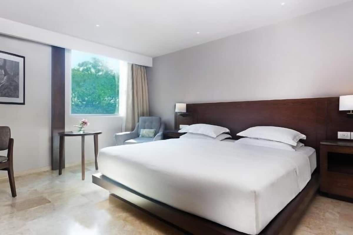 A modern hotel room with a large bed, a chair, a small table with a vase, and a window with curtains.