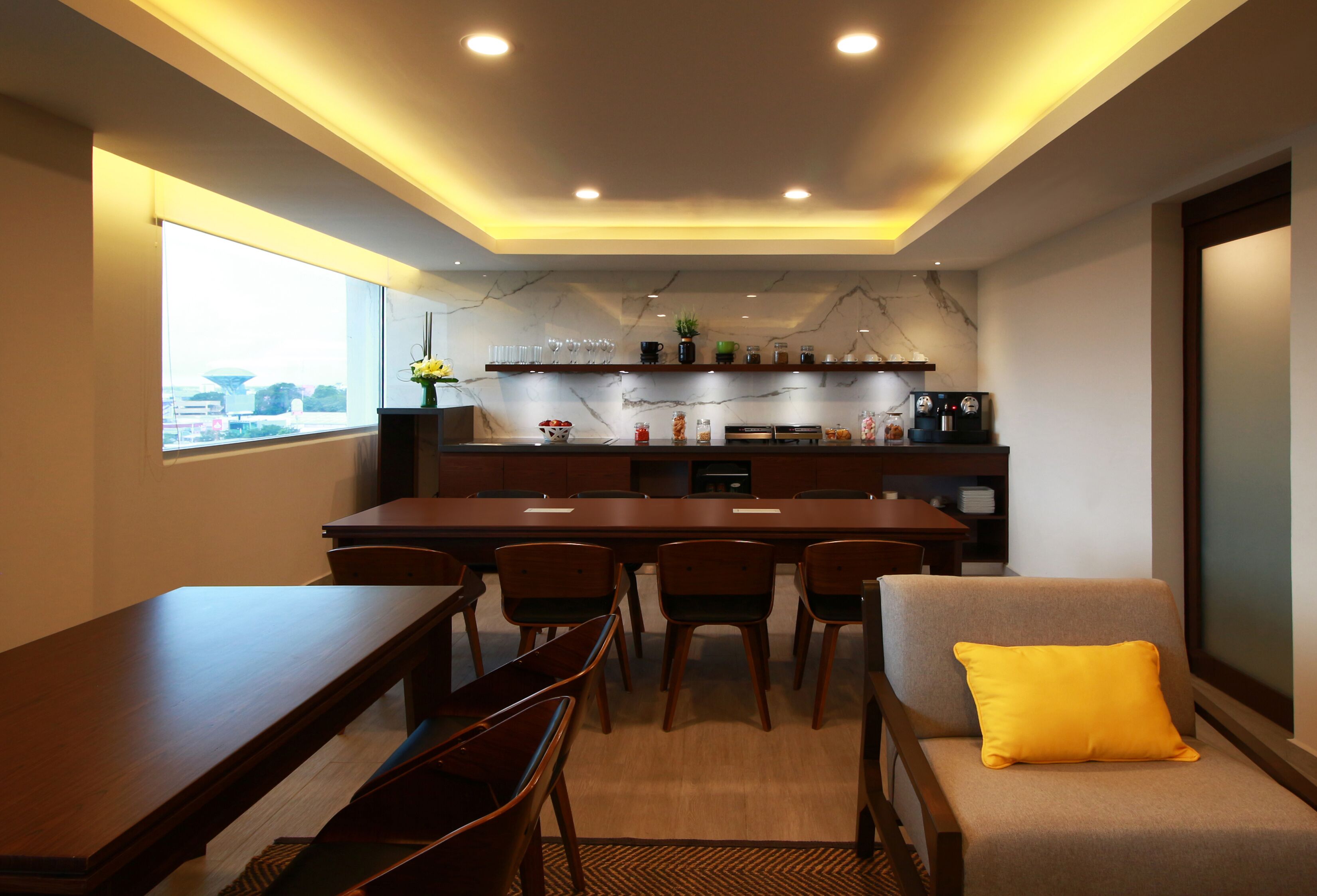 Un espace repas moderne avec une longue table, des chaises et un canapé agrémenté d’un coussin jaune.