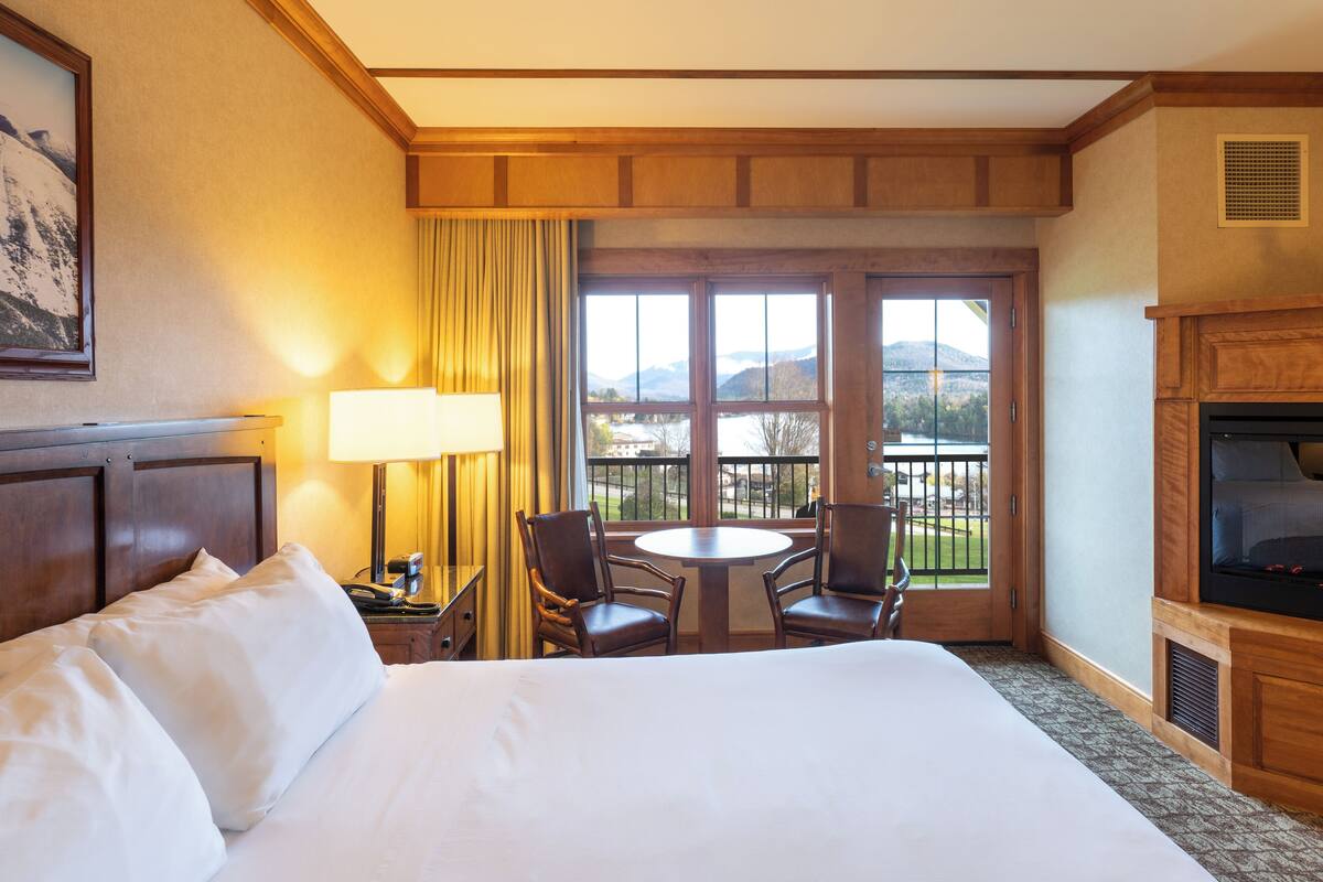Une chambre d’hôtel avec un grand lit, un bureau avec une lampe, deux chaises, une petite table, un téléviseur et une fenêtre donnant sur les montagnes.