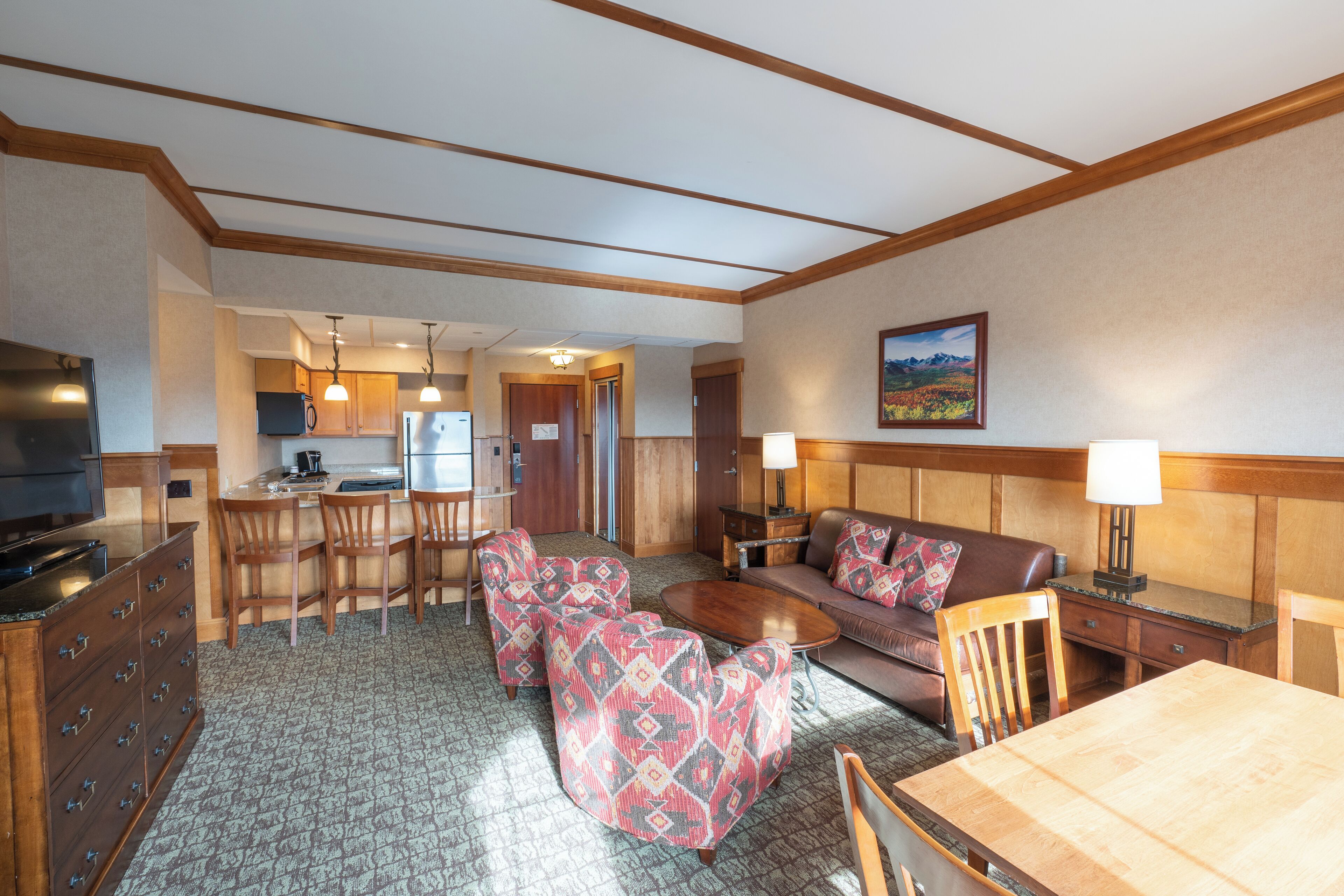 A hotel room with a kitchen area, a dining table, a sofa, and a TV.