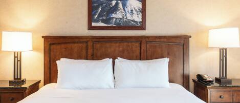 A hotel room with a large bed, two bedside lamps, a wooden headboard, and a framed mountain landscape picture on the wall.
