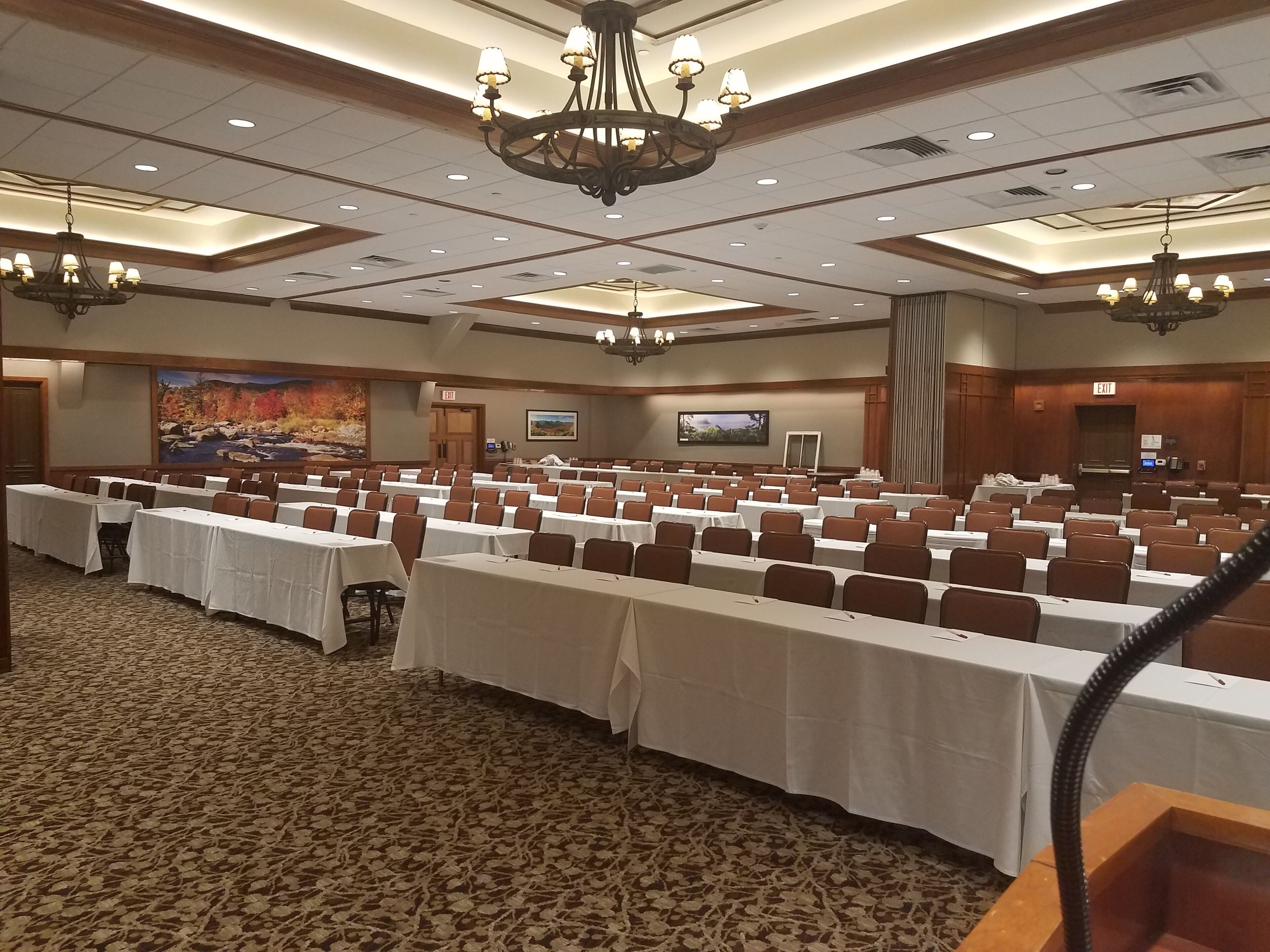 A large conference room with rows of tables and chairs, a chandelier, and a large screen on the wall.
