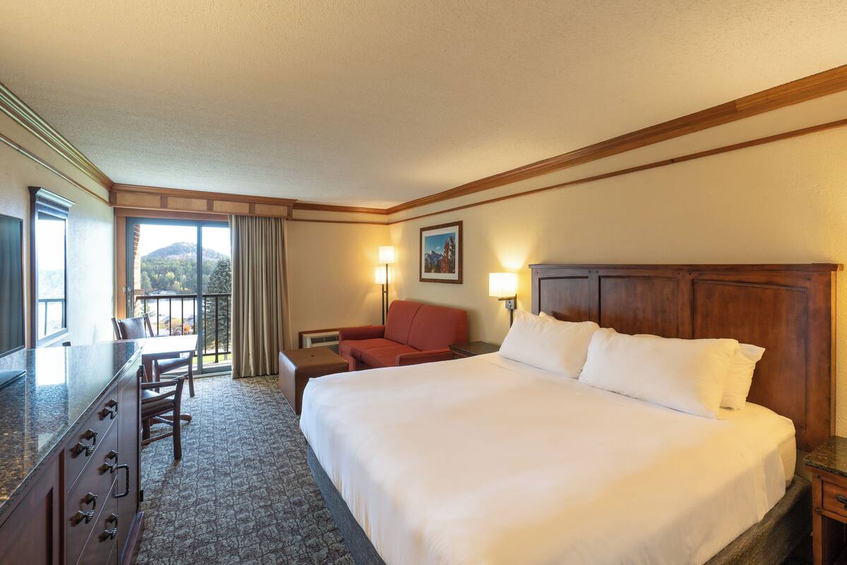 Une chambre d’hôtel avec un grand lit, un bureau avec des tiroirs, une chaise, une lampe et une fenêtre donnant sur une vue sur la montagne.