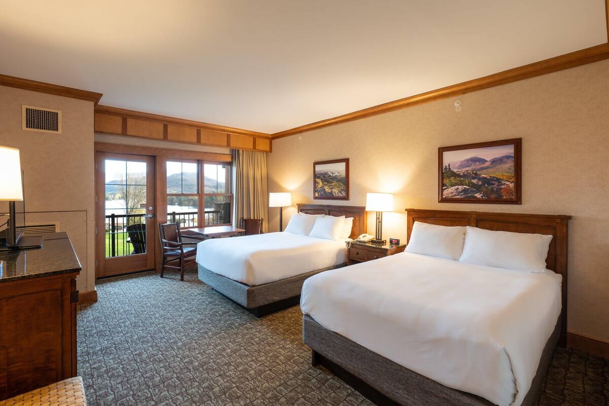 Une chambre d’hôtel avec deux lits, un bureau, une chaise et un balcon offrant une vue sur les montagnes.