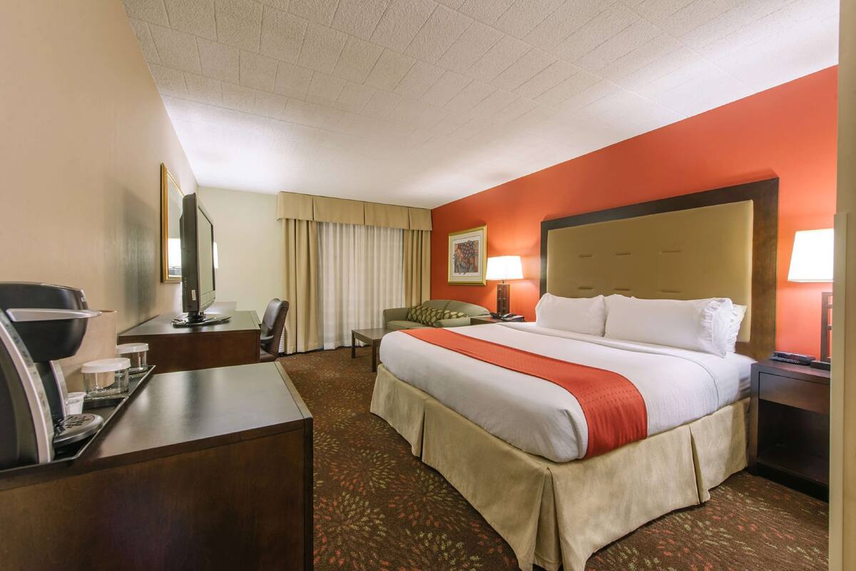 A hotel room with a large bed, a desk with a coffee maker, a chair, a nightstand with a lamp, and a window with curtains.