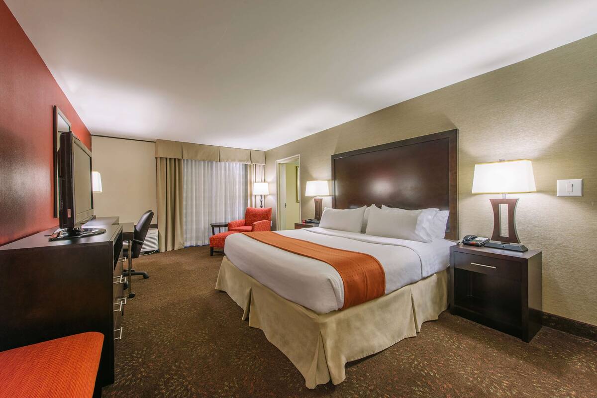 A hotel room with a large bed, a desk, a chair, a nightstand, and a lamp.