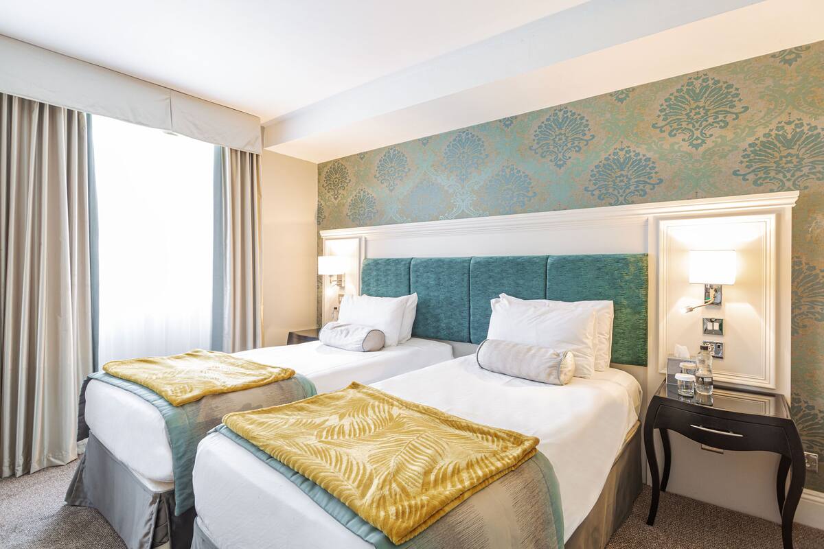 A hotel room with two beds, a nightstand with a lamp, a mirror, and a patterned wall.