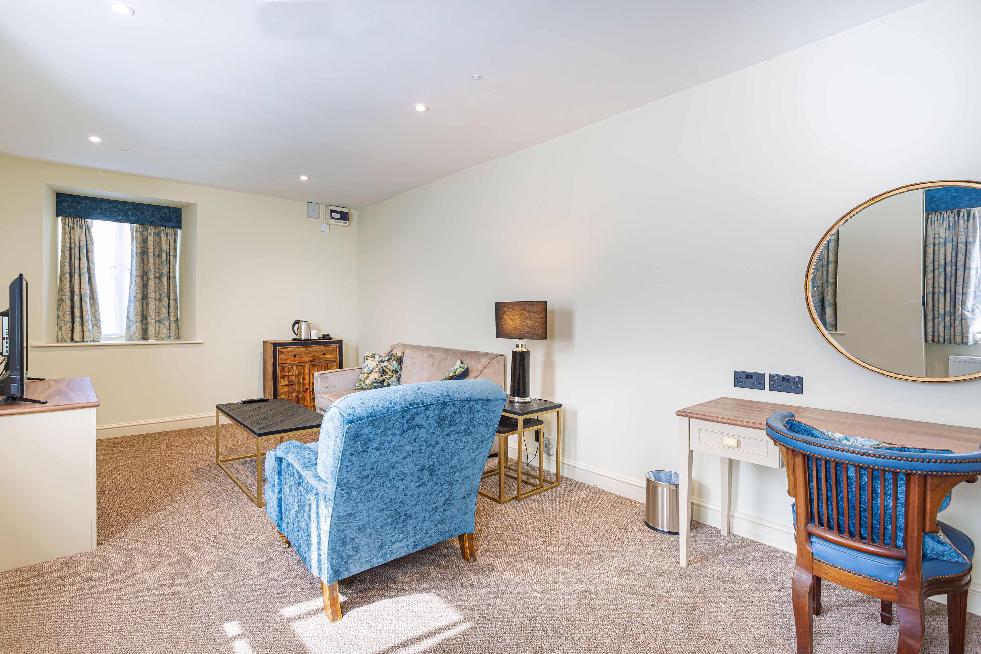 A room with a blue armchair, a wooden desk, a mirror, a lamp, and a window with curtains.