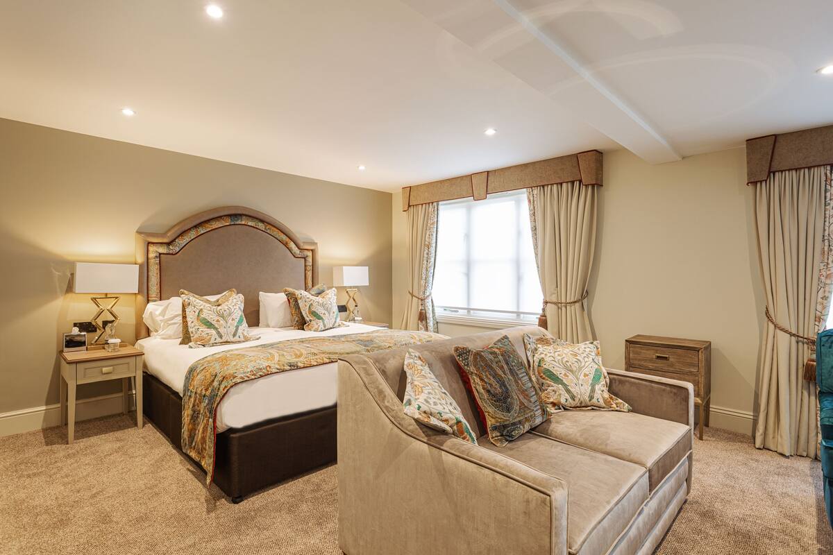 A hotel room with a large bed, a sofa, a nightstand, a lamp, and a window with curtains.