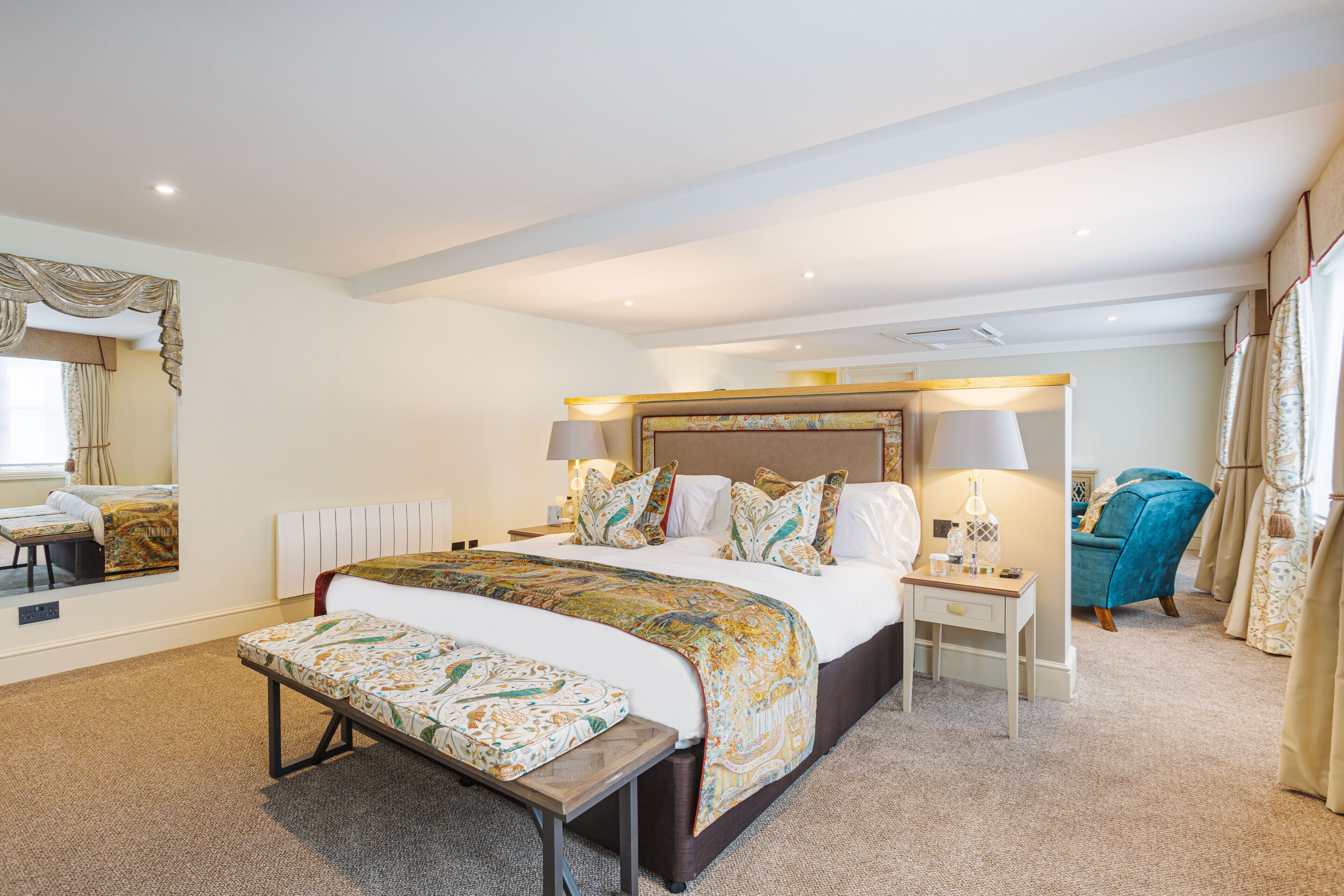 A hotel room with a large bed, bedside tables, a bench, a mirror, and a window with curtains.