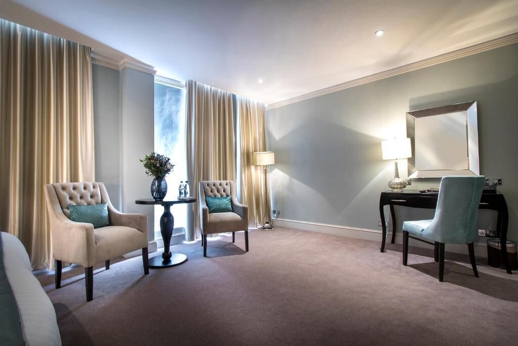 Quarto de hotel com uma janela grande, duas poltronas, uma mesa de trabalho com luminária e um espelho.
