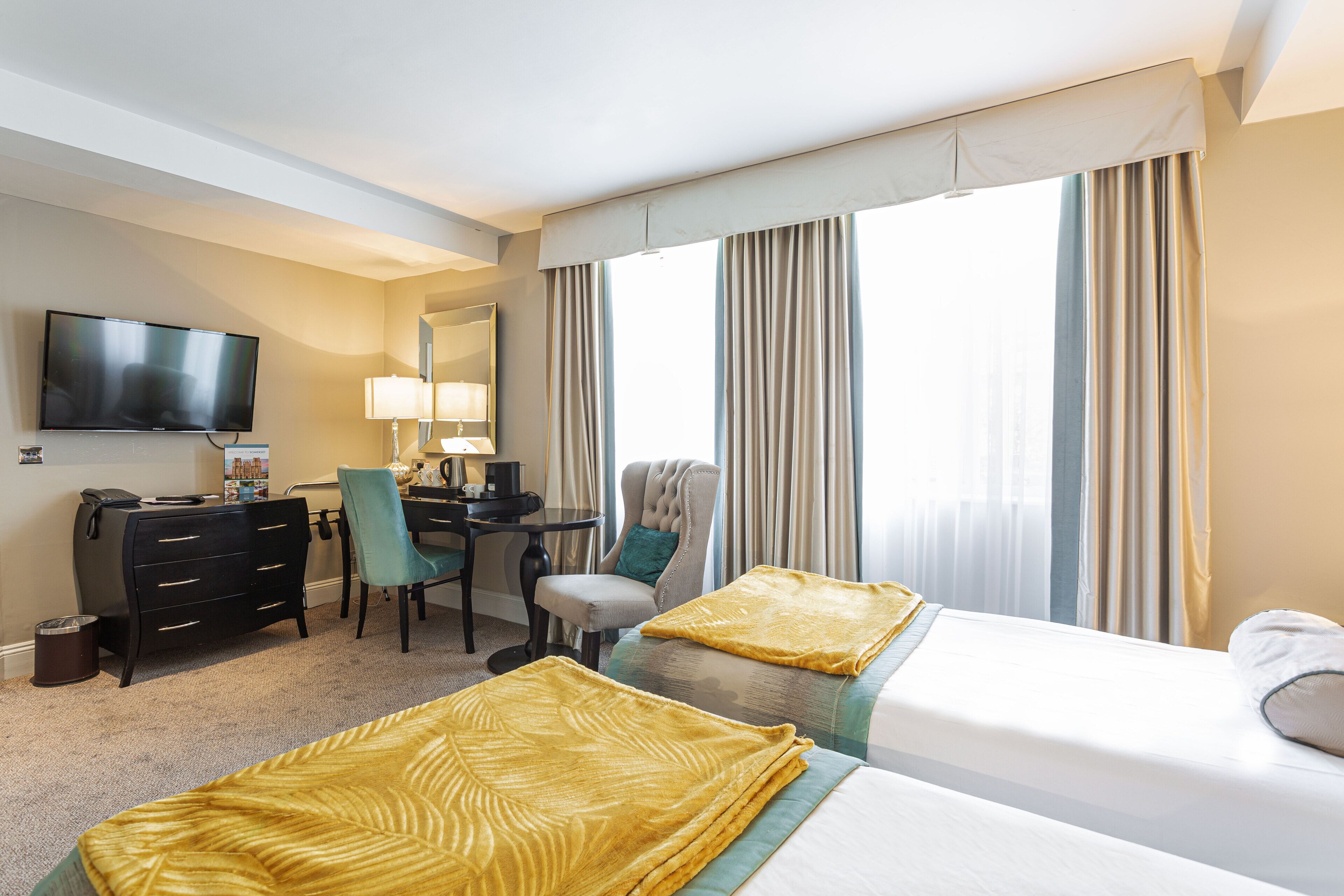 A hotel room with two beds, a desk, a chair, a TV, and a large window with curtains.