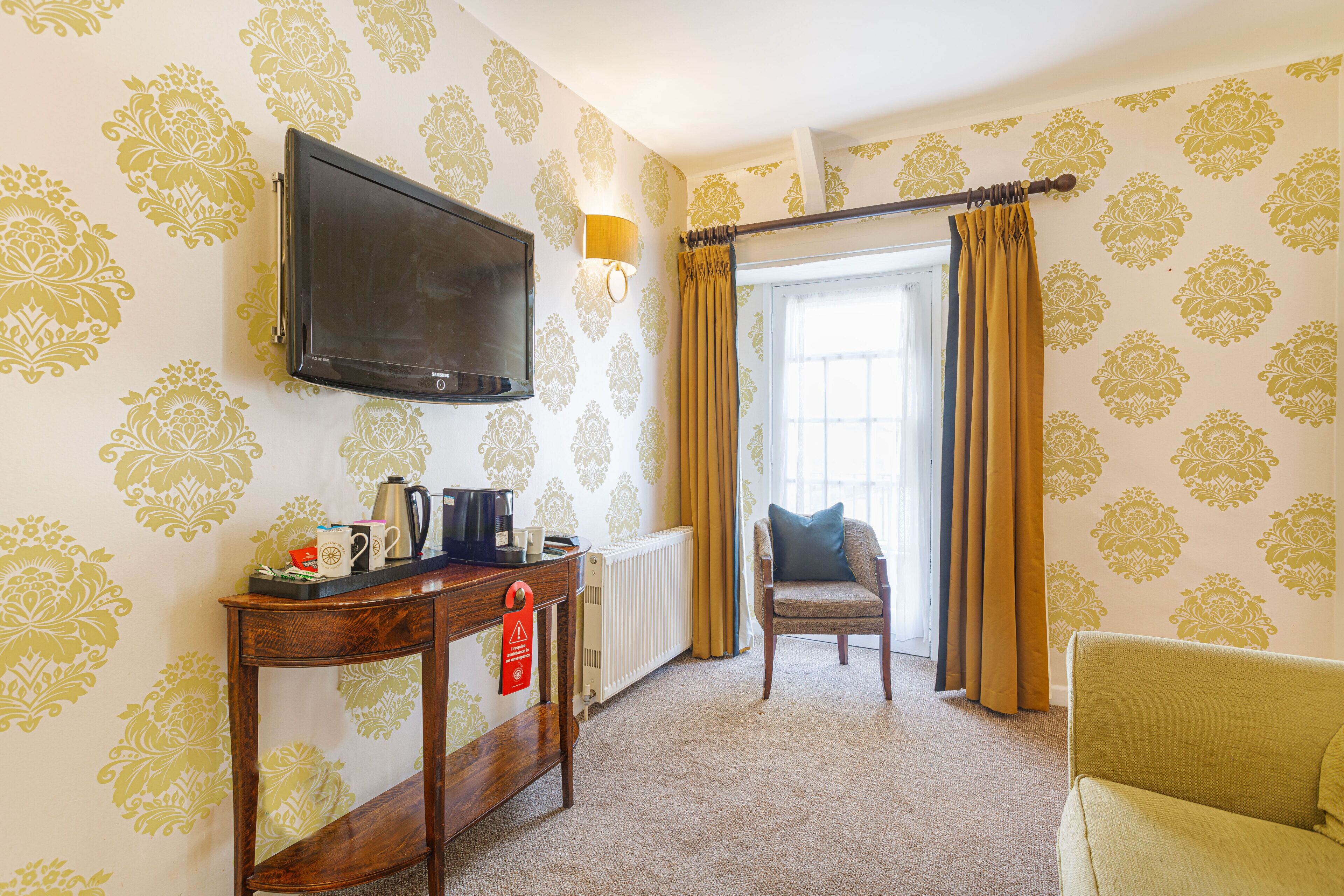 A hotel room with a television, a table with a kettle, a chair, and a sofa.