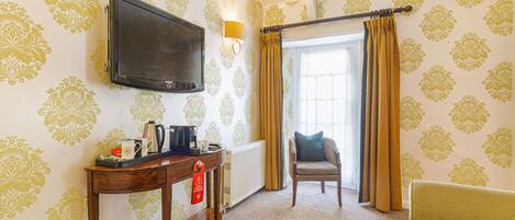 A hotel room with a television, a table with a kettle, a chair, and a sofa.