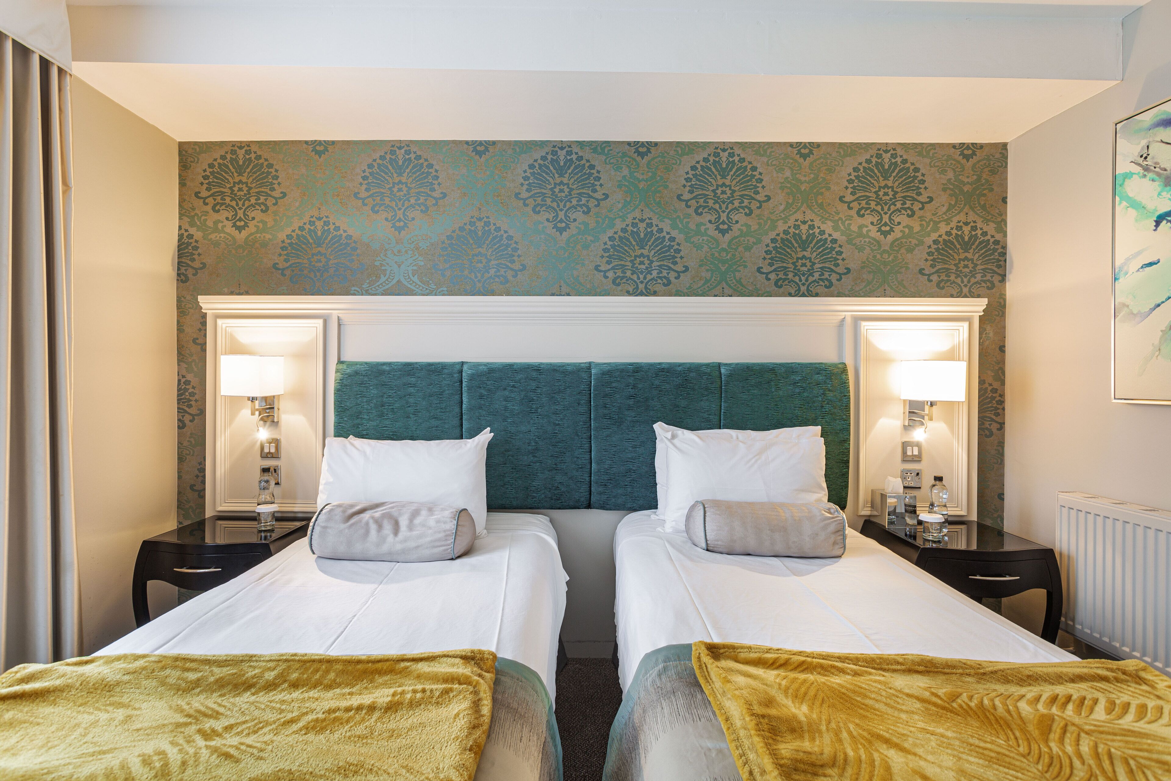 A hotel room with two beds, a green headboard, bedside lamps, and a patterned wallpaper.