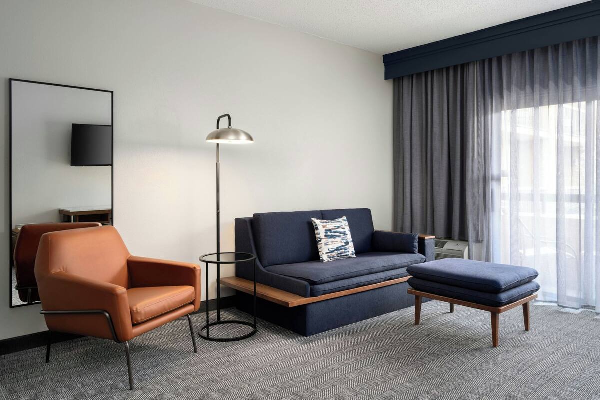 Una habitación de hotel moderna con sofá, sillón, reposapiés, lámpara de pie y un espejo grande.