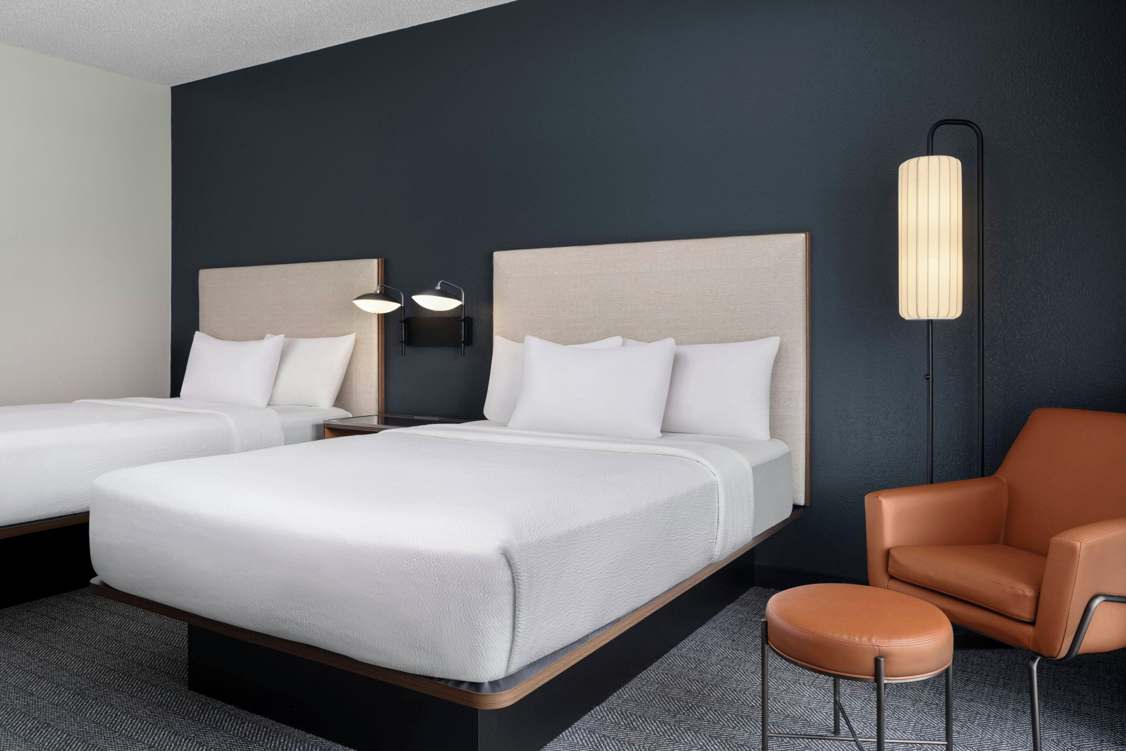 A hotel room with two beds, a brown leather chair, a small stool, and a tall floor lamp.