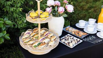 A buffet table with various food items, including sandwiches, pastries, and a fruit basket, set against a backdrop of greenery.
