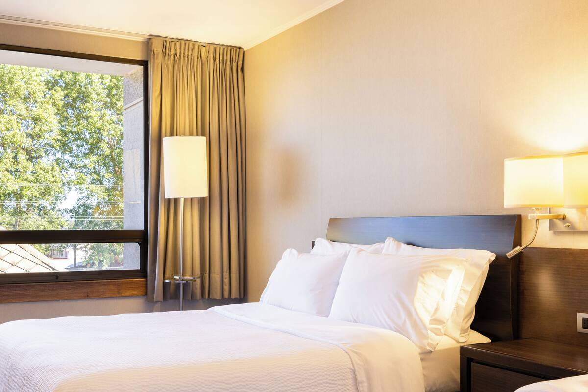 Une chambre d’hôtel avec un grand lit, deux lampes de chevet, une fenêtre avec des rideaux et une vue sur les arbres à l’extérieur.