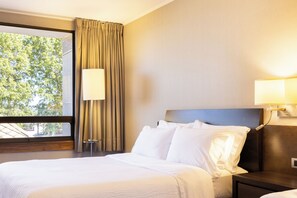 Habitación de hotel con una cama grande, dos lámparas de noche, una ventana con cortinas y vistas a árboles.