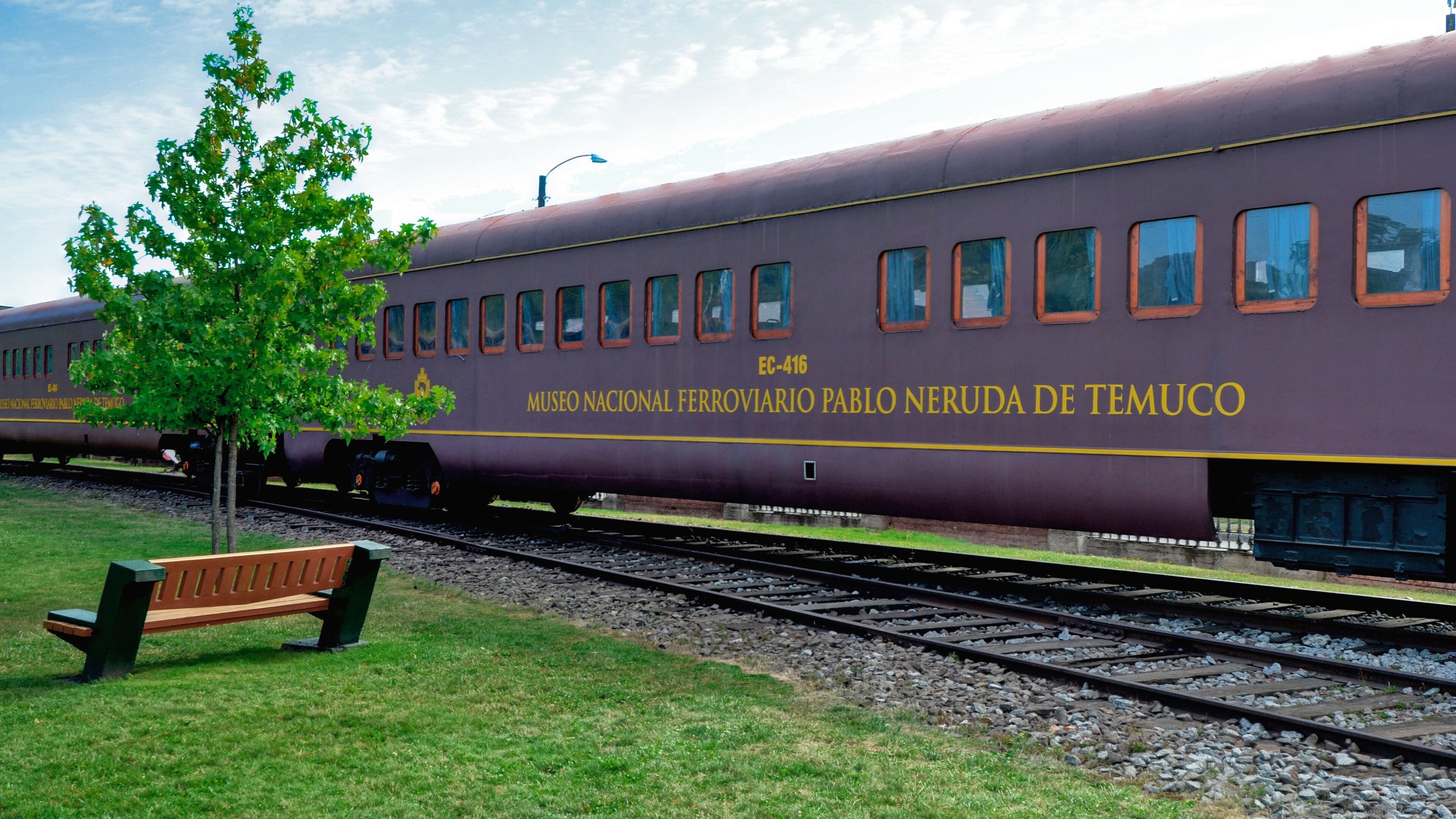 A train with "MUSEO NACIONAL FERROVIARIO PABLO NERUDA DE TEMUCO" written on its side, parked on tracks with a bench and a tree nearby.