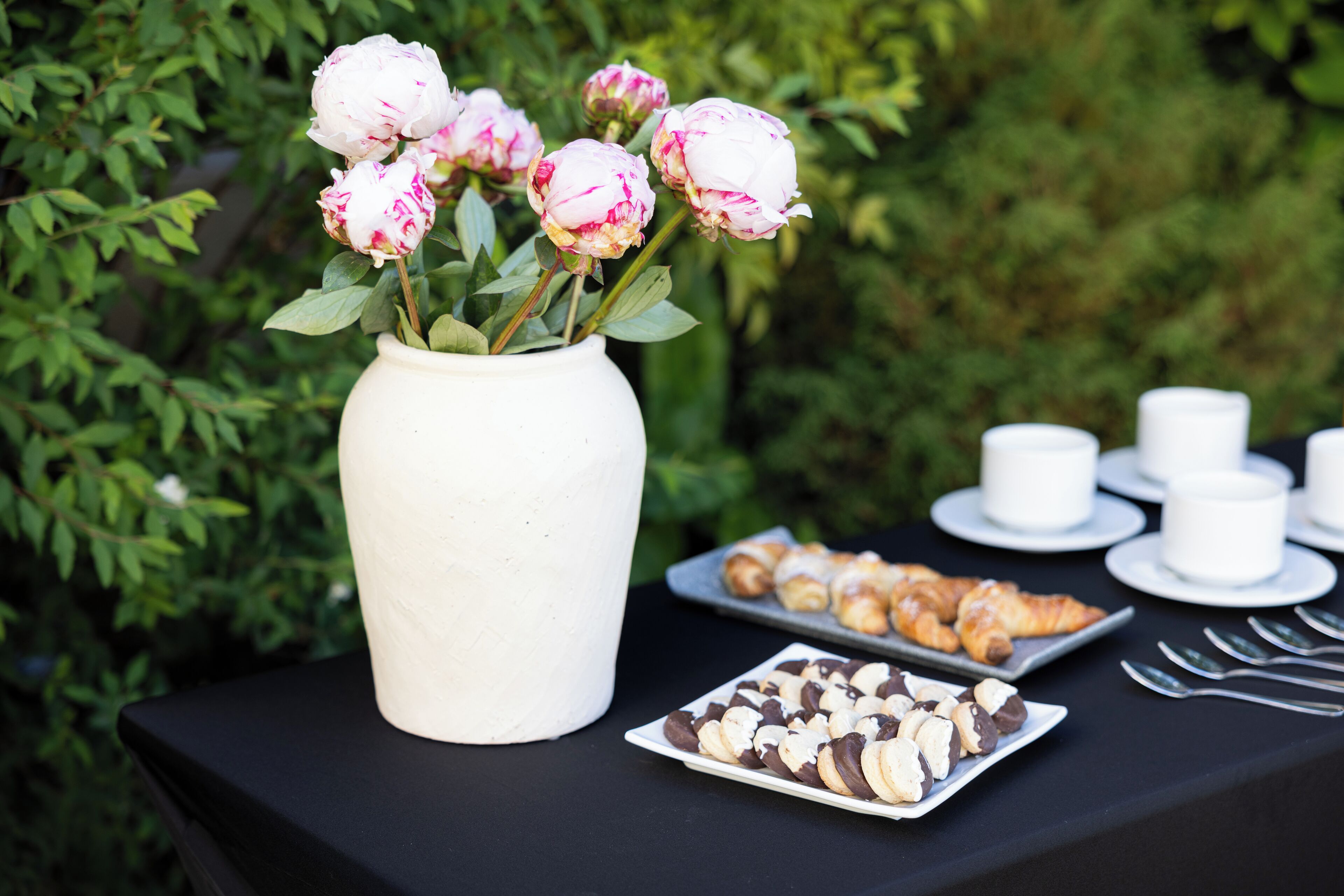 A table with a white vase of peonies, a plate of pastries, and white cups and saucers.