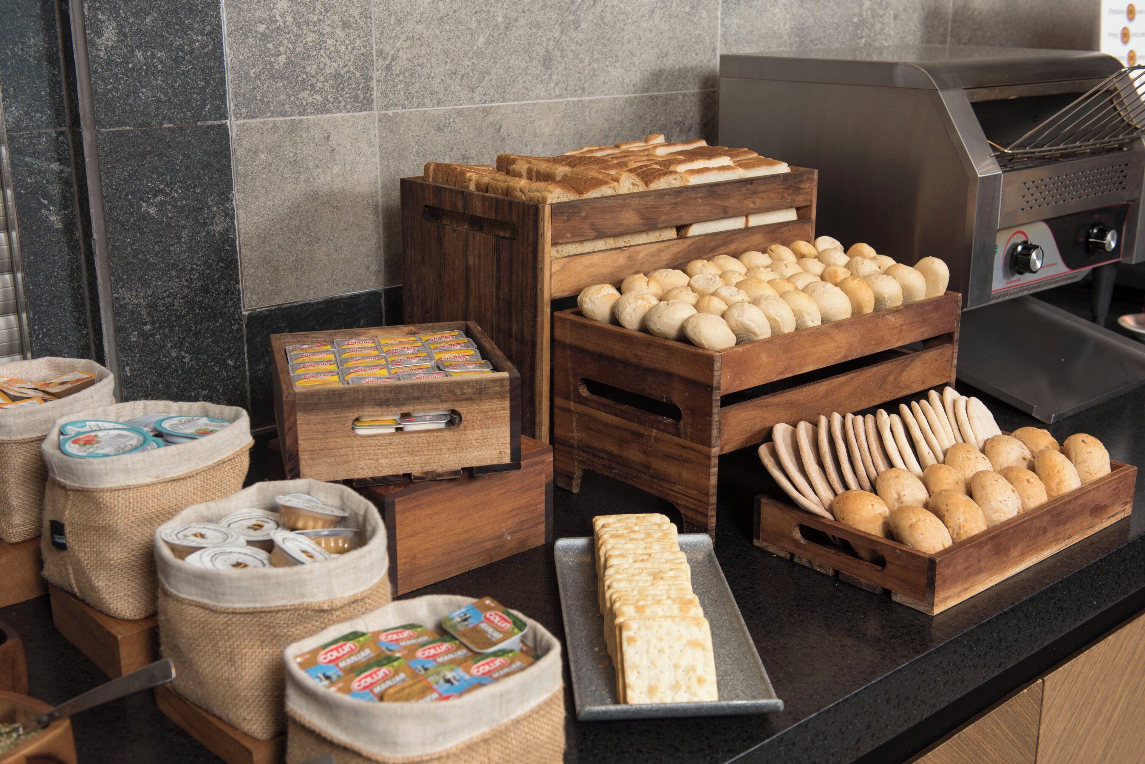 Uma bancada de cozinha com diversos pães e produtos de panificação expostos em caixas e cestos de madeira.