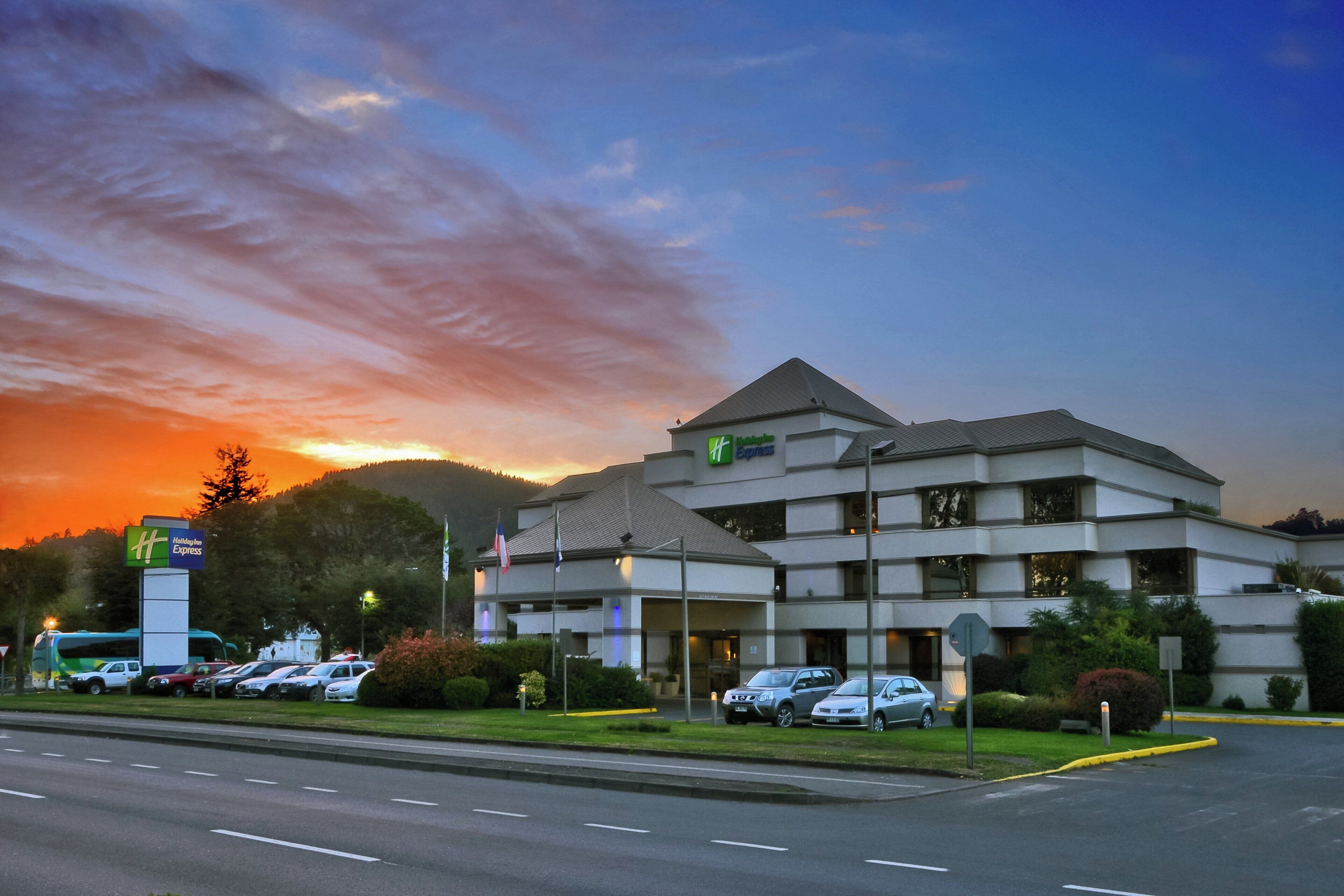 Un hotel de varios pisos con un llamativo letrero de Holiday Inn Express, estacionamiento y un paisaje montañoso de fondo.
