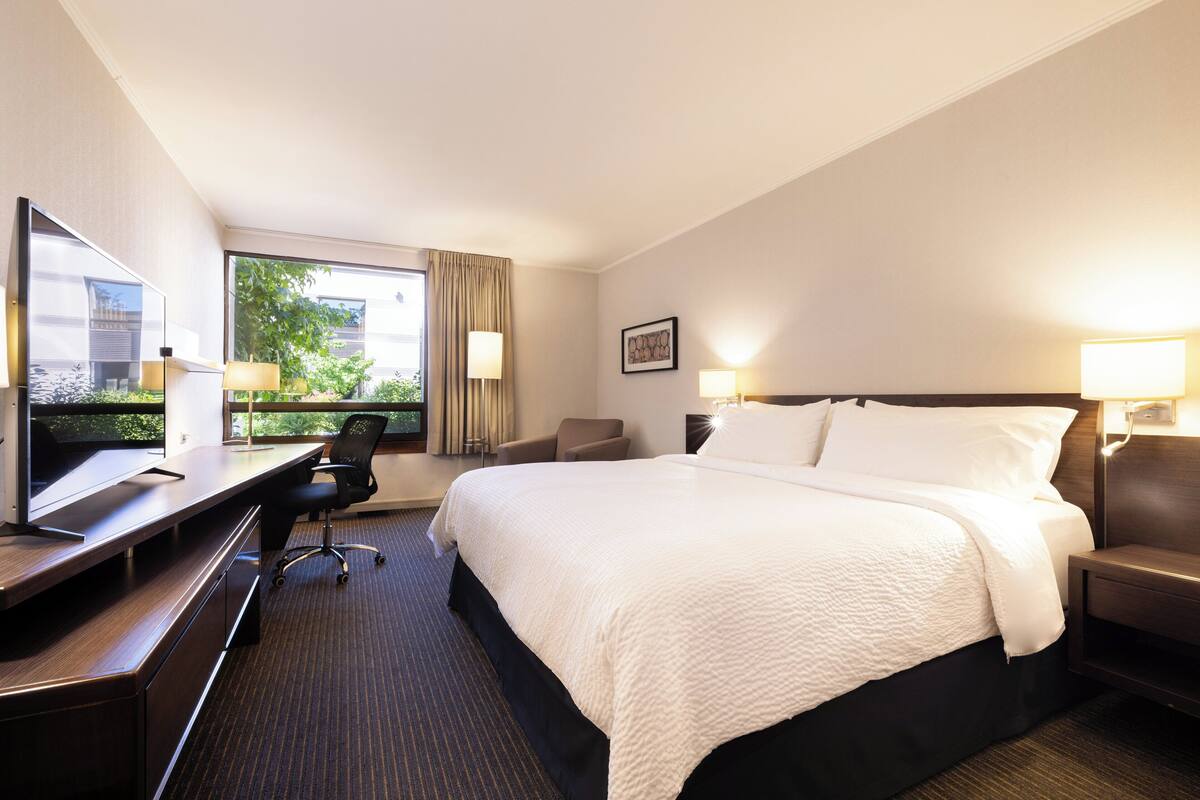Une chambre d’hôtel avec un grand lit, un bureau avec une chaise, une télévision et une fenêtre avec des rideaux.